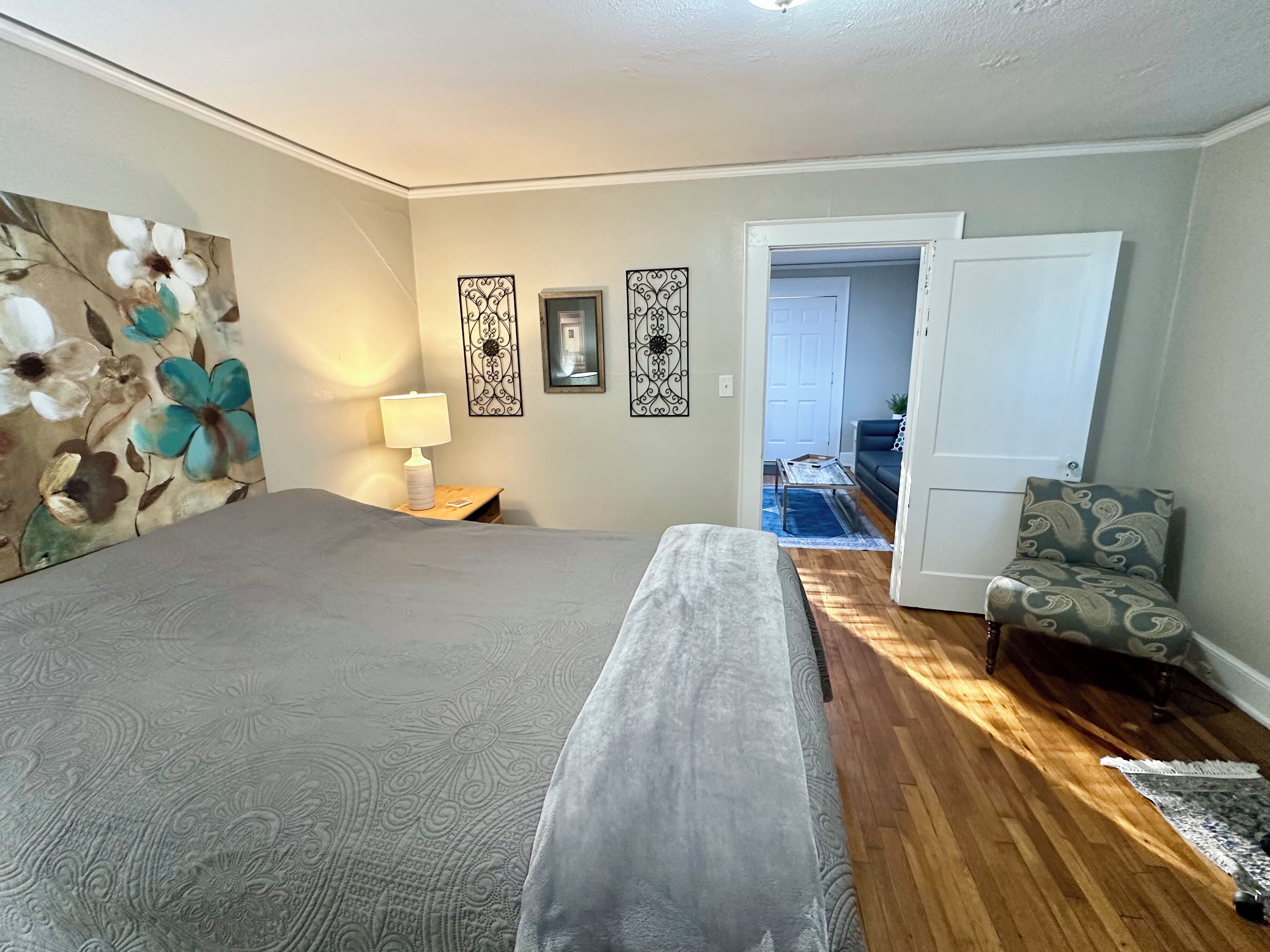 Une chambre à coucher avec un grand lit, une table de chevet avec une lampe, une chaise et une porte donnant sur une autre pièce.