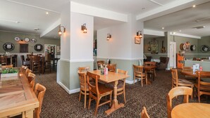 A spacious dining area with wooden tables and chairs, a kitchen in the background, and wall-mounted clocks. - Wren's Nest Manorcombe 41 (Gunnislake)
