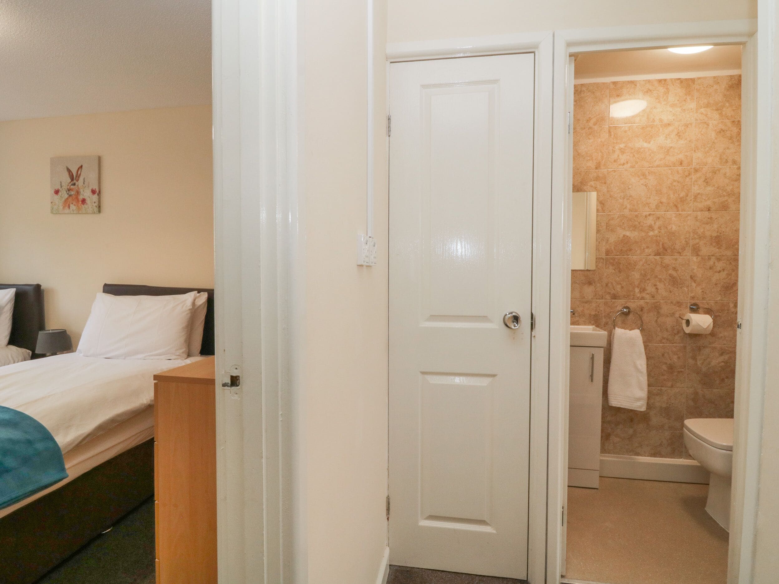 A hotel room with a bed, a nightstand, a painting on the wall, and a bathroom with a toilet, sink, and towel rack.