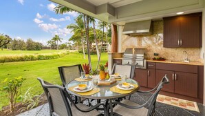 A dining area with a view of a golf course, a kitchen with a grill, and a dining table set with plates and glasses.