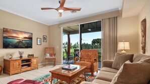 A living room with a sofa, armchairs, a coffee table, and a television. There is a ceiling fan, a window with curtains, and a view of the outdoors.