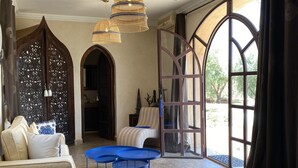 Ein geräumiges Wohnzimmer mit einem beigefarbenen Sofa, einem gestreiften Sofa und einem blauen Couchtisch. Drei hängende Korbtablellen und ein großes Bogenfenster mit Vorhängen.