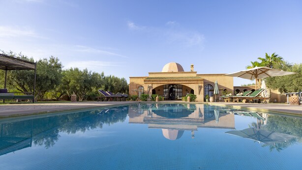 Ein Swimmingpool mit klarer Spiegelung, ein Gebäude mit Kuppel und Bögen sowie Außenbereich mit Sonnenschirmen.