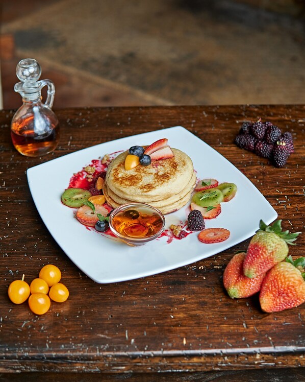 A square white plate with pancakes topped with berries and a small bowl of syrup, surrounded by fresh fruits and a bottle of syrup on a wooden table. - Estancia San Juan (Sayausí)