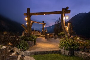 Un espacio de estar al aire libre rústico con estructuras de madera y mesas de piedra, rodeado de follaje y montañas al fondo.