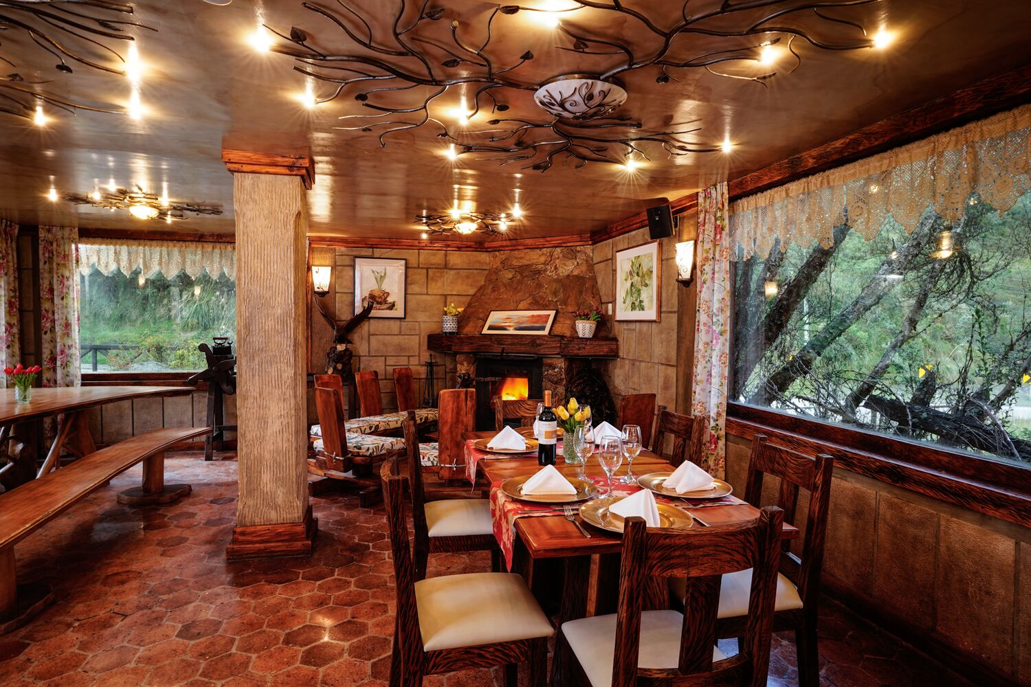 Zona pranzo rustica con camino in pietra, mobili in legno e vista sugli alberi attraverso un'ampia finestra.