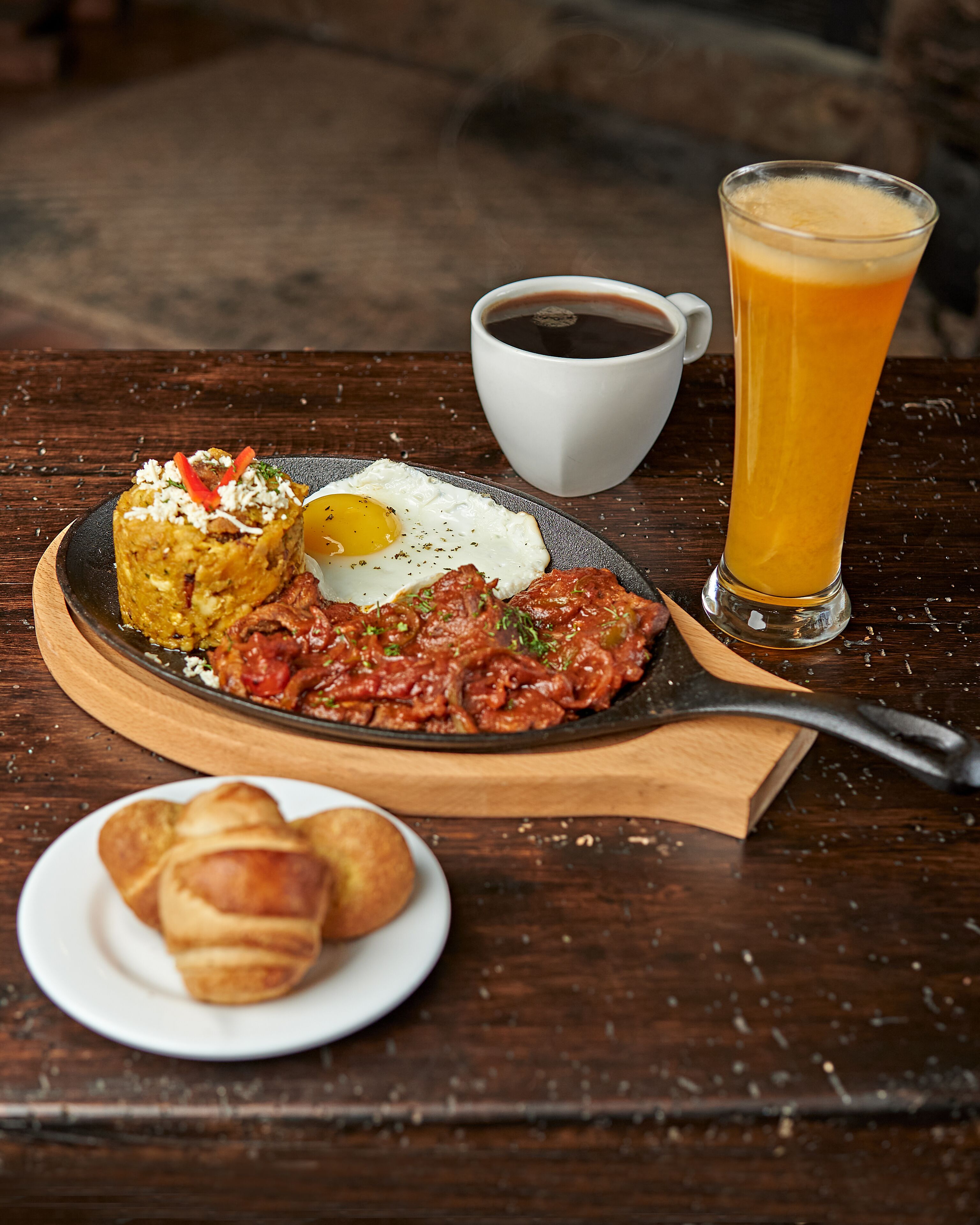 Ein Frühstücksteller mit Eiern, Kartoffelrösti und Wurst, ein Glas Orangensaft und eine Tasse Kaffee auf einem Holztisch.