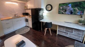 Een moderne keuken met witte kasten, een zwarte koelkast, een magnetron en een houten aanrecht. Er is een zithoek met een witte tafel en twee stoelen. Een klok en een televisie hangen aan de muur.