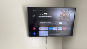 A flat-screen TV mounted on a wall displaying a YouTube video. Below the TV, there is a grey filing cabinet with multiple drawers and a black trash bin. - Twin Oaks Extended Stay One Bedroom - 2817 Apt 6 (Syracuse)