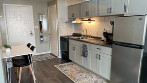 A modern kitchen with grey cabinets, a stainless steel refrigerator, a stove, and a sink. There is a small dining area with a table and chairs. - Twin Oaks Extended Stay One Bedroom - 2817 Apt 6 (Syracuse)