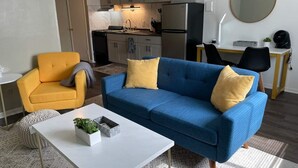 A modern living room with a blue sofa, yellow cushions, a white coffee table with a plant, a yellow armchair, and a kitchen in the background. - Twin Oaks Extended Stay One Bedroom - 2817 Apt 6 (Syracuse)