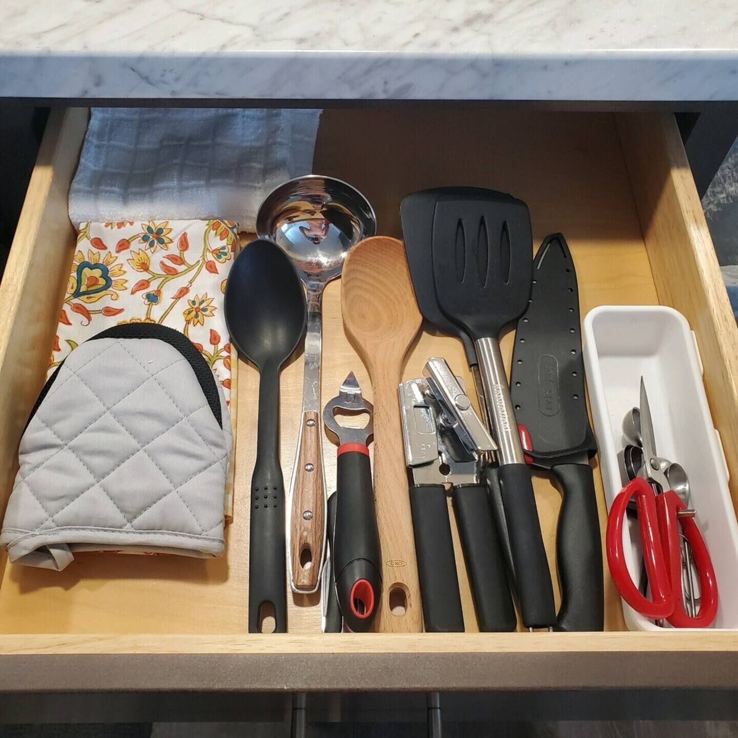 A drawer with various kitchen utensils including a wooden spoon, a black spatula, and a knife.
