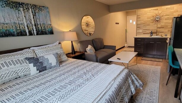A modern hotel room with a large bed, a sofa, a small table, and a kitchenette area.