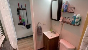 Une petite salle de bain comprenant des toilettes, un lavabo et un rideau de douche. On y trouve des produits de nettoyage sur une étagère et un porte-serviettes.