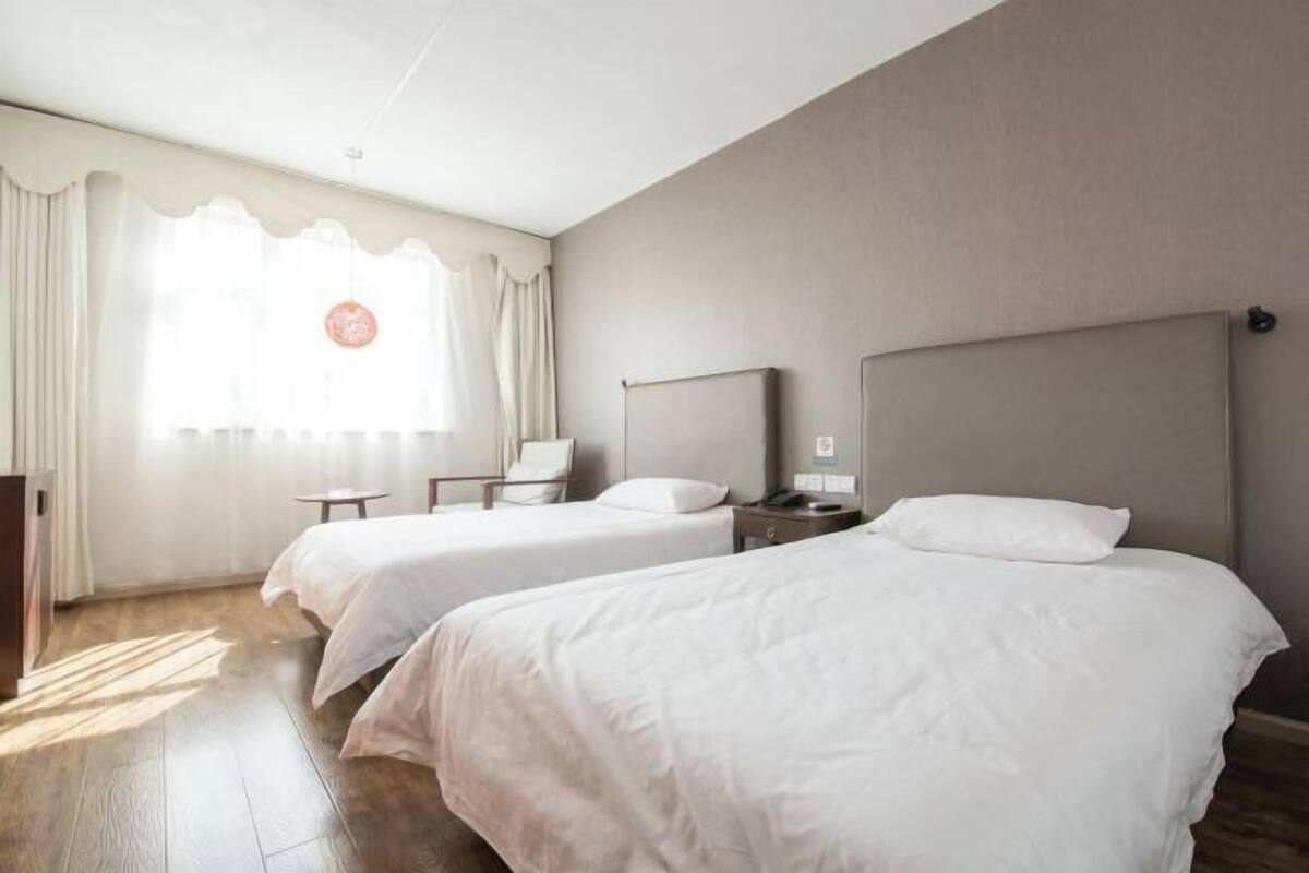 Quarto de hotel com duas camas, uma escrivaninha, uma cadeira, uma mesa de cabeceira e uma janela com cortinas.