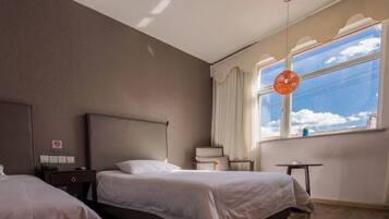Ein Hotelzimmer mit zwei Betten, einem Schreibtisch, einem Stuhl und einem Fenster mit Blick zum Himmel.