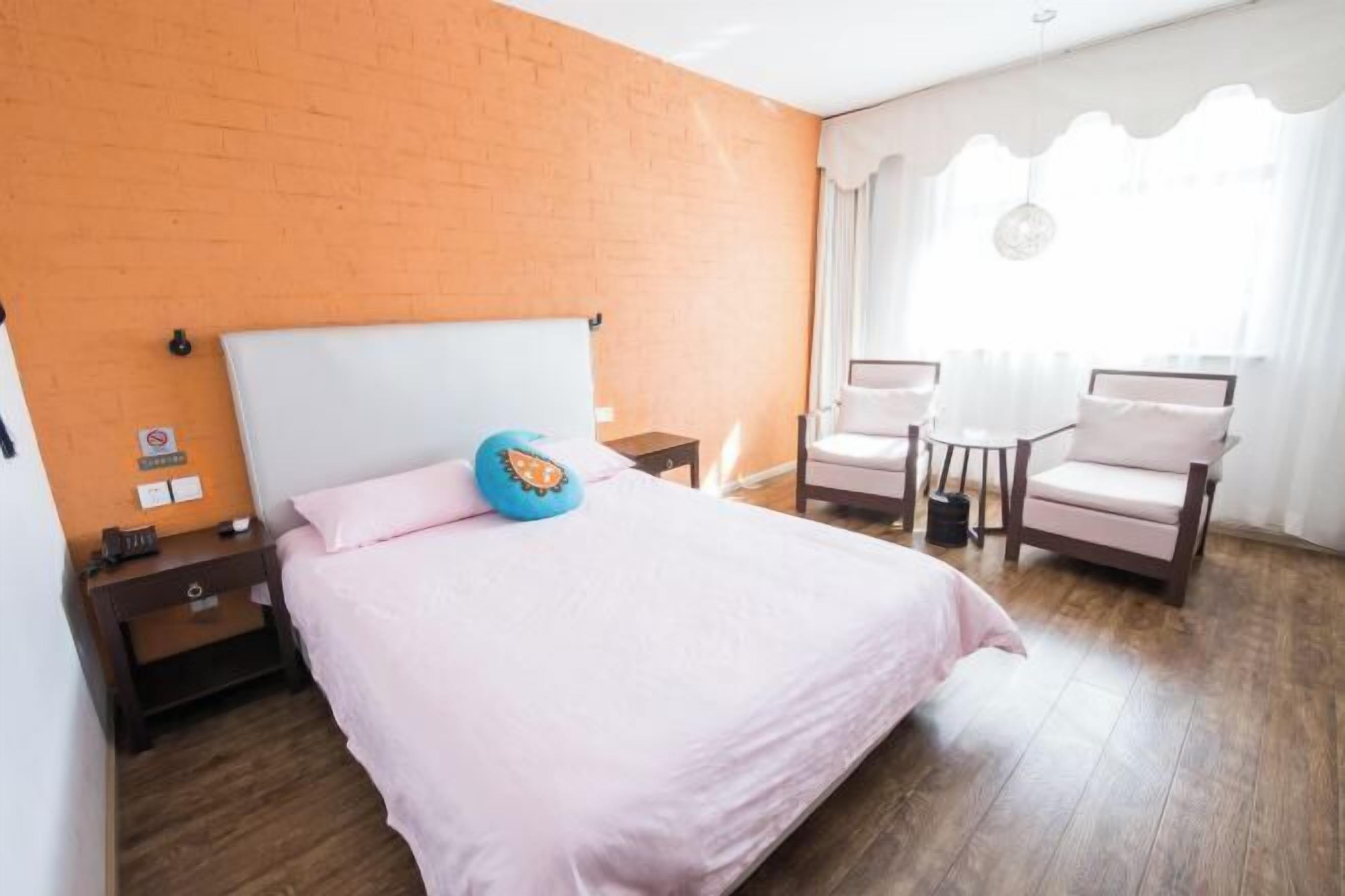 A hotel room with a brick wall, a bed with a white headboard, a nightstand, a desk, a chair, and a window with curtains.