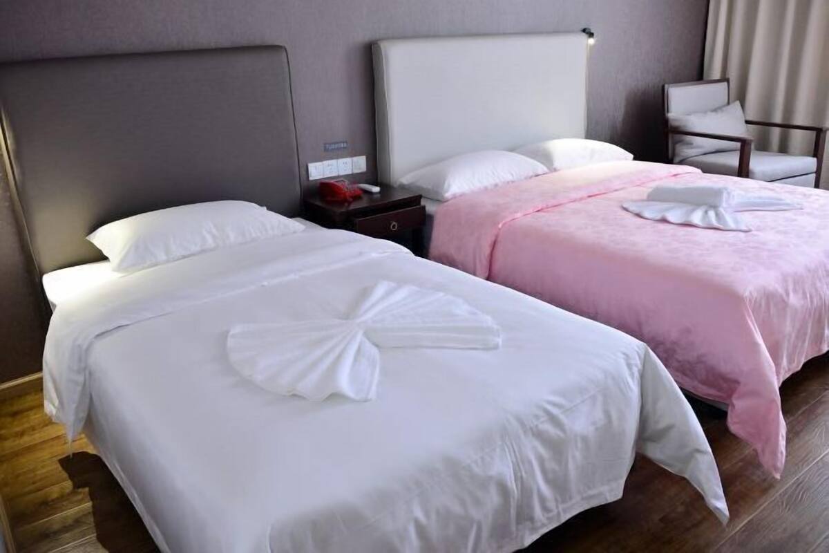 Quarto de hotel com duas camas, cada uma com almofada e toalha dobrada. Há uma cadeira e uma mesinha de cabeceira com abajur.