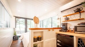 A compact kitchen with a wooden countertop, a microwave, a stove, and a small refrigerator. There is a window with blinds, a plant, and a bed with a white blanket in the adjacent room. - Moss Oak - Container Home Near Magnolia and Baylor (Waco)
