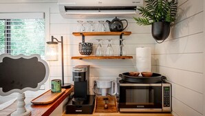 A modern kitchen with a coffee maker, microwave, and shelves holding glassware and a kettle. - Moss Oak - Container Home Near Magnolia and Baylor (Waco)