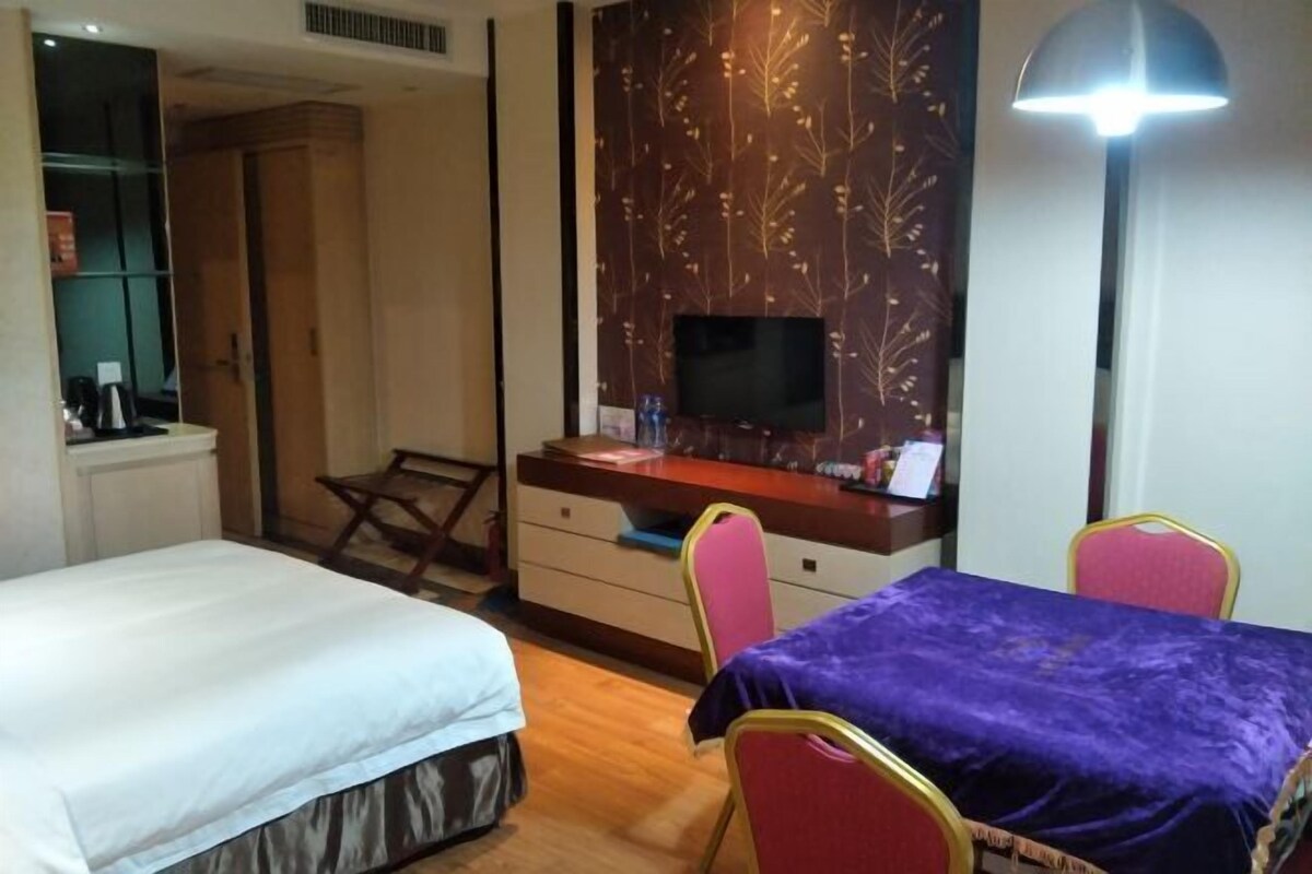A hotel room with a bed, a desk with a TV, a table with chairs, and a lamp.
