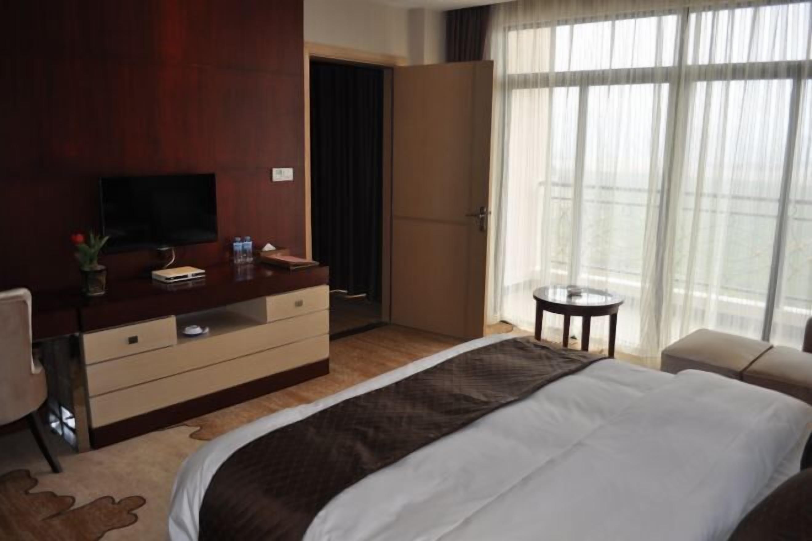 Habitación de hotel con una cama grande, un escritorio con televisor, una mesita pequeña y una ventana con cortinas.