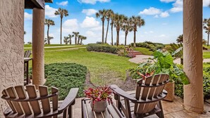 A covered patio with wooden chairs and a potted plant, overlooking a well-maintained lawn and palm trees.
