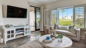 A modern living room with a flat-screen TV, a white entertainment unit, a beige sofa with cushions, a round coffee table with books and a plant, and large windows with a view of a lawn and palm trees.