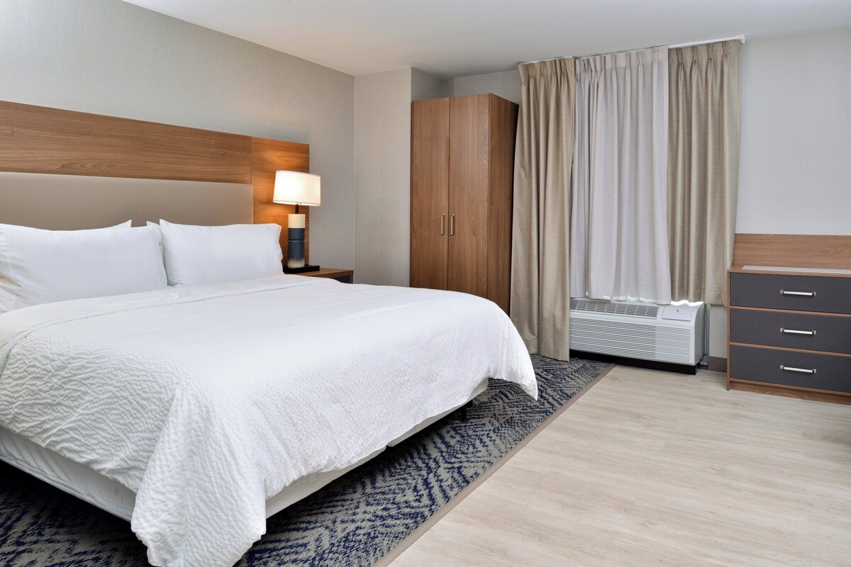 A hotel room with a large bed, a wooden headboard, a nightstand with a lamp, a wardrobe, and a window with curtains.