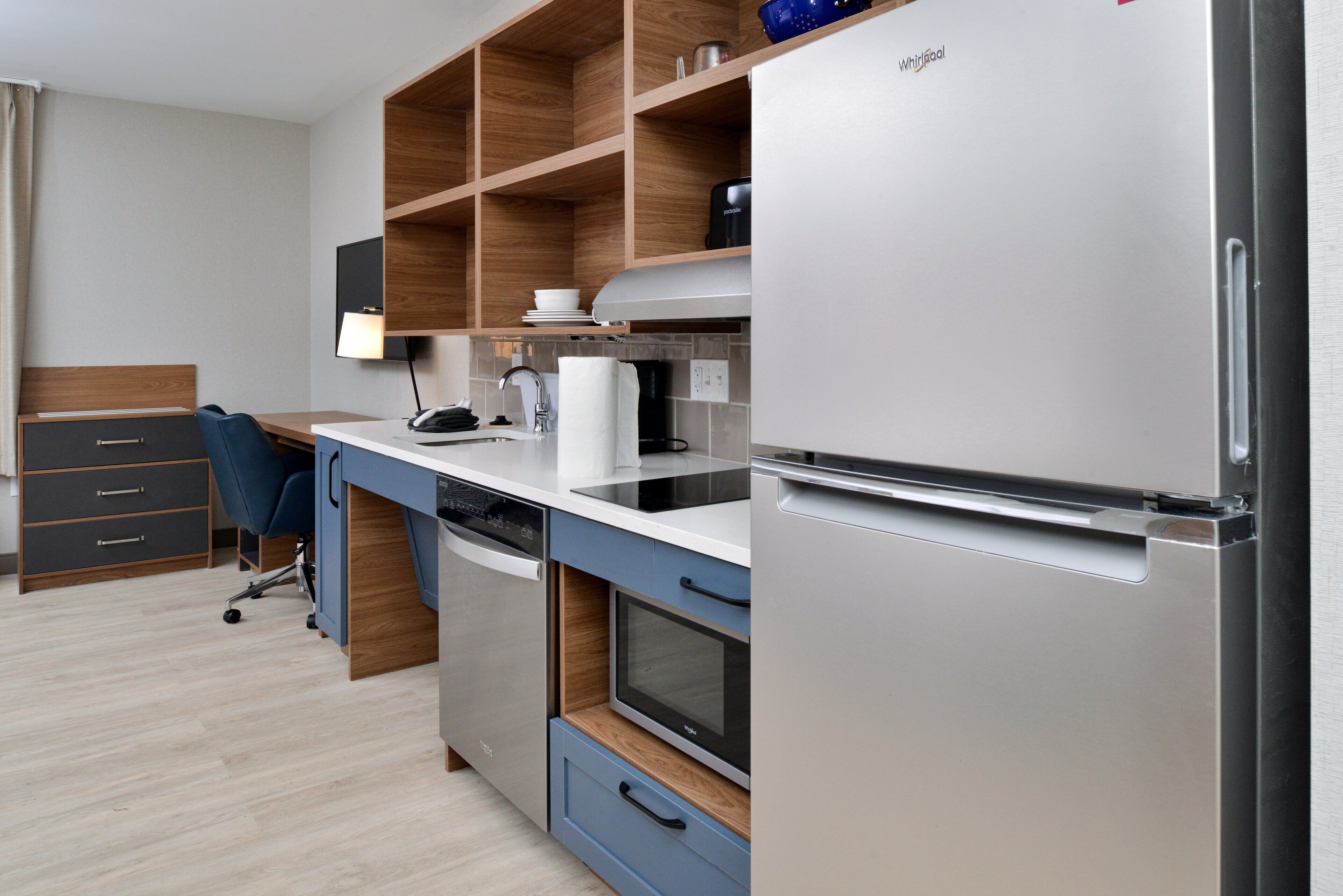 A modern kitchen with a built-in dishwasher, microwave, and refrigerator. There is a wooden shelf above the counter and a blue chair in the corner.