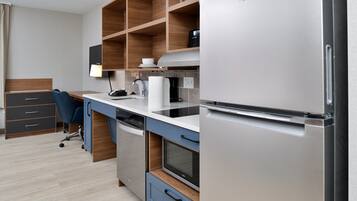 A modern kitchen with a built-in dishwasher, microwave, and refrigerator. There is a wooden shelf above the counter and a blue chair in the corner.