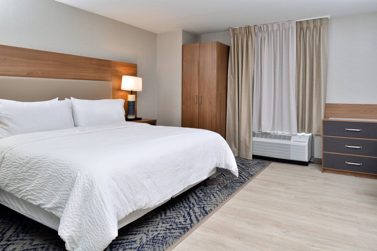 A hotel room with a large bed, a wooden headboard, a nightstand with a lamp, a wardrobe, and a window with curtains.