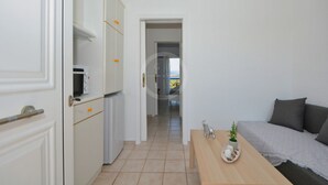 En smal hall leder in till ett sovrum med en säng, ett litet bord med en växt och en mikrovågsugn i köksdelen.