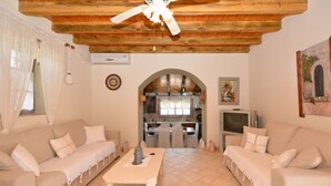 A living room with two beige sofas, a white coffee table, a ceiling fan, a kitchen area with a TV, and a painting on the wall. - Estia Stone-Built Cottage, the perfect family getaway (Argolida)