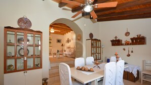 A dining area with a table set for four, white chairs, a ceiling fan, and a view into another room with a sofa and a lamp. - Estia Stone-Built Cottage, the perfect family getaway (Argolida)