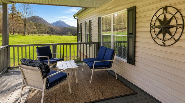 Une véranda abritée avec deux chaises et une table, une décoration murale représentant une boussole, et une vue sur un champ verdoyant et des montagnes.