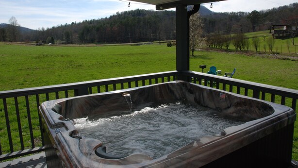 A hot tub on a covered deck with a view of a green field and trees in the background. - Blue Ridge Cozy Cabin with Full Mountain View (Blue Ridge)