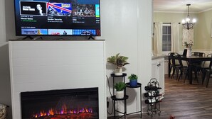 A living room with a fireplace, a TV mounted on the wall, a dining area with a table and chairs, and a hallway with a chandelier. - Blue Ridge Cozy Cabin with Full Mountain View (Blue Ridge)