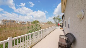 Ein Balkon mit weißer Reling, einem Korbstuhl und Blick auf ein Feld mit hohem Gras und Häusern im Hintergrund.