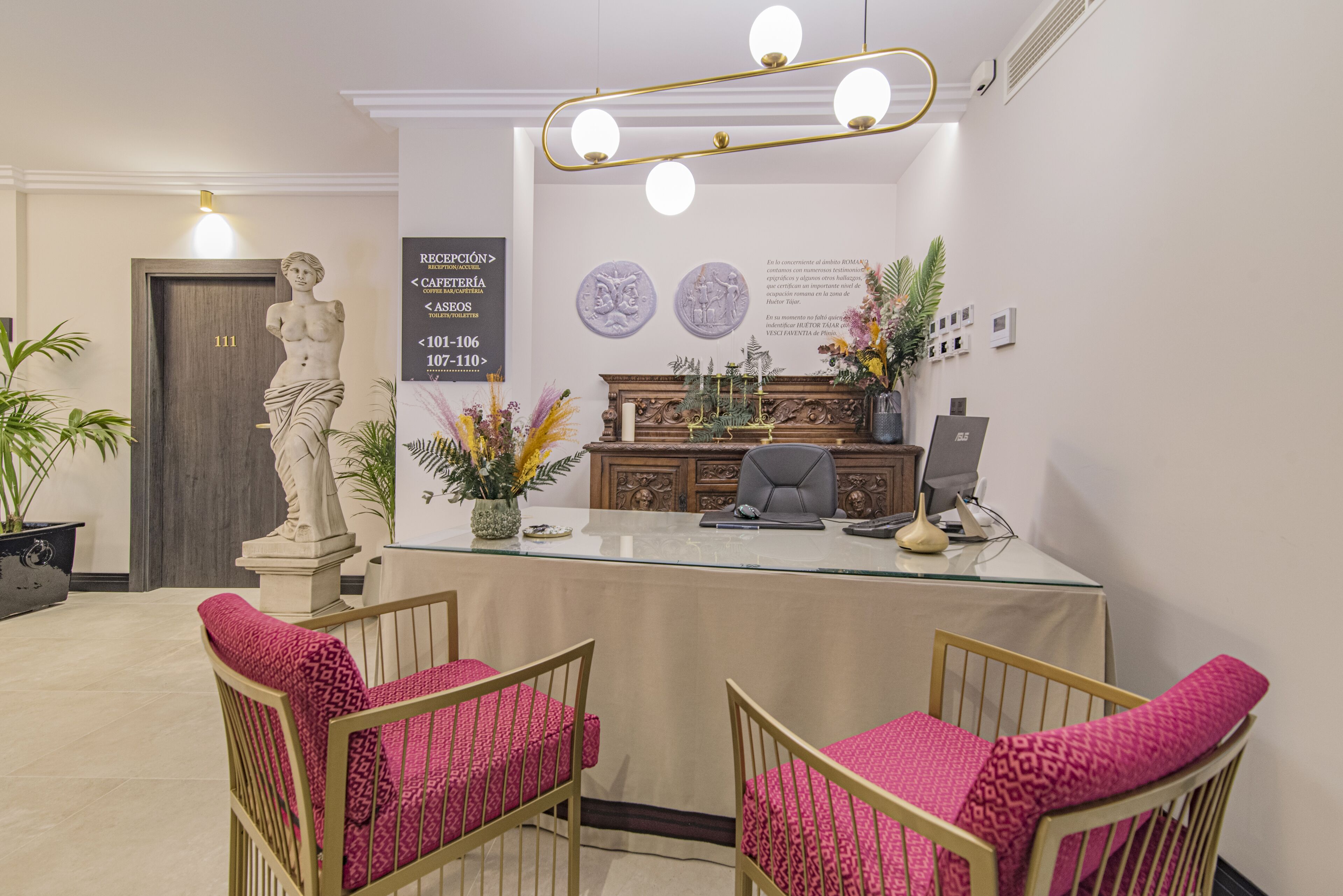 Un área de recepción con una estatua de mármol, un mostrador de recepción, dos sillas con cojines rosa y un aparador decorativo.