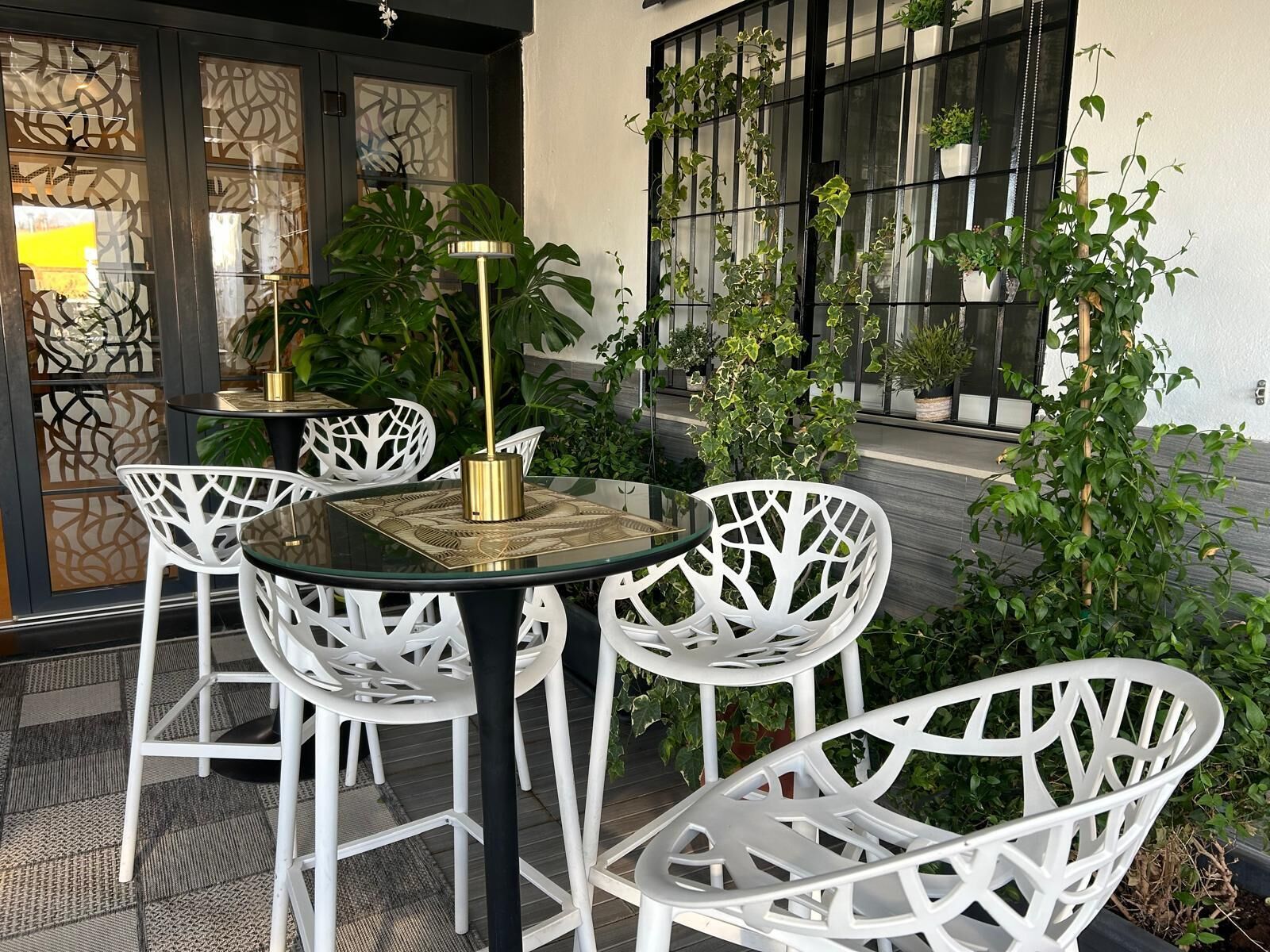 An outdoor seating area with white chairs and a glass table, surrounded by potted plants and a decorative wall.