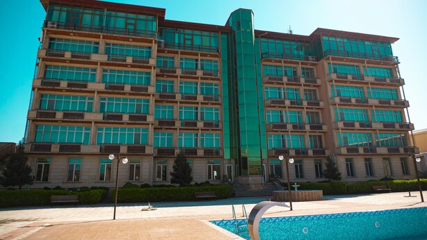 A multi-story hotel with a central glass elevator shaft, balconies on each floor, and a swimming pool in the foreground. - Olympic Hotel & Resort (Absheron)