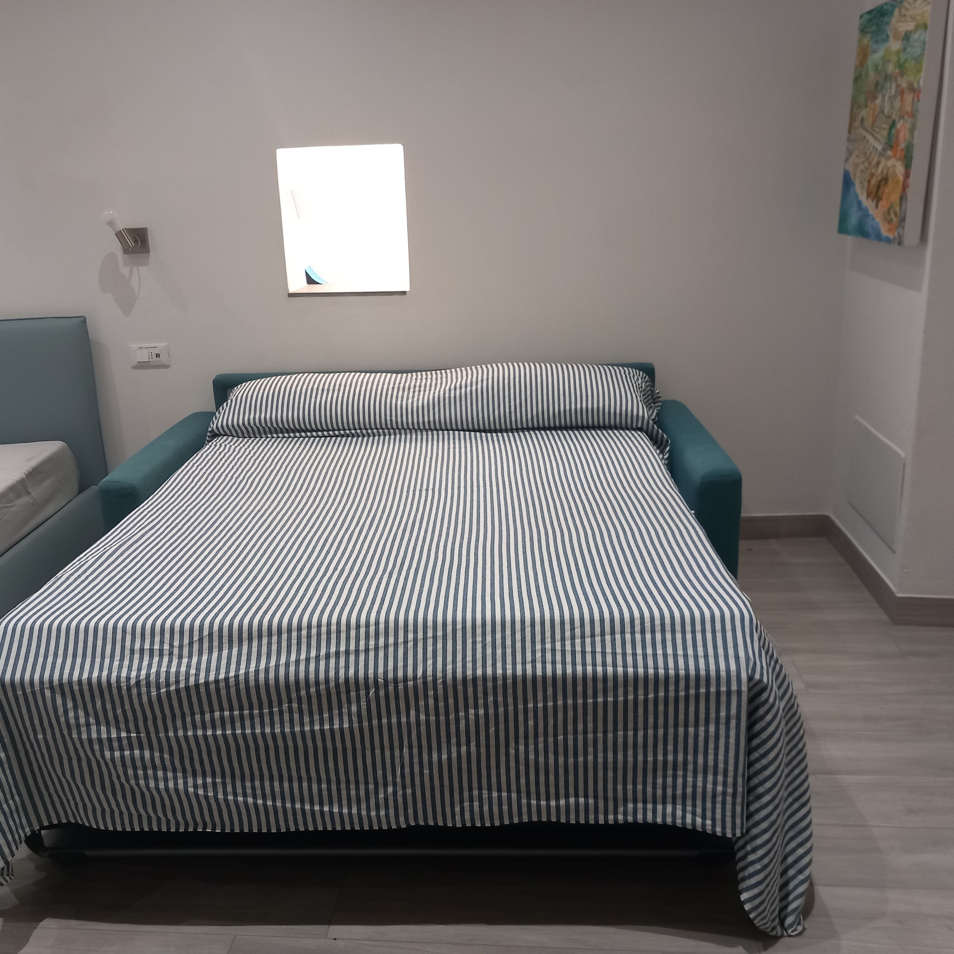 Un lit avec une couvre-lit à rayures, une petite fenêtre et un luminaire fixé au mur.