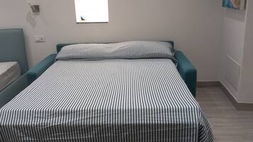 Una cama con una colcha a rayas, una ventana pequeña y una lámpara de pared.