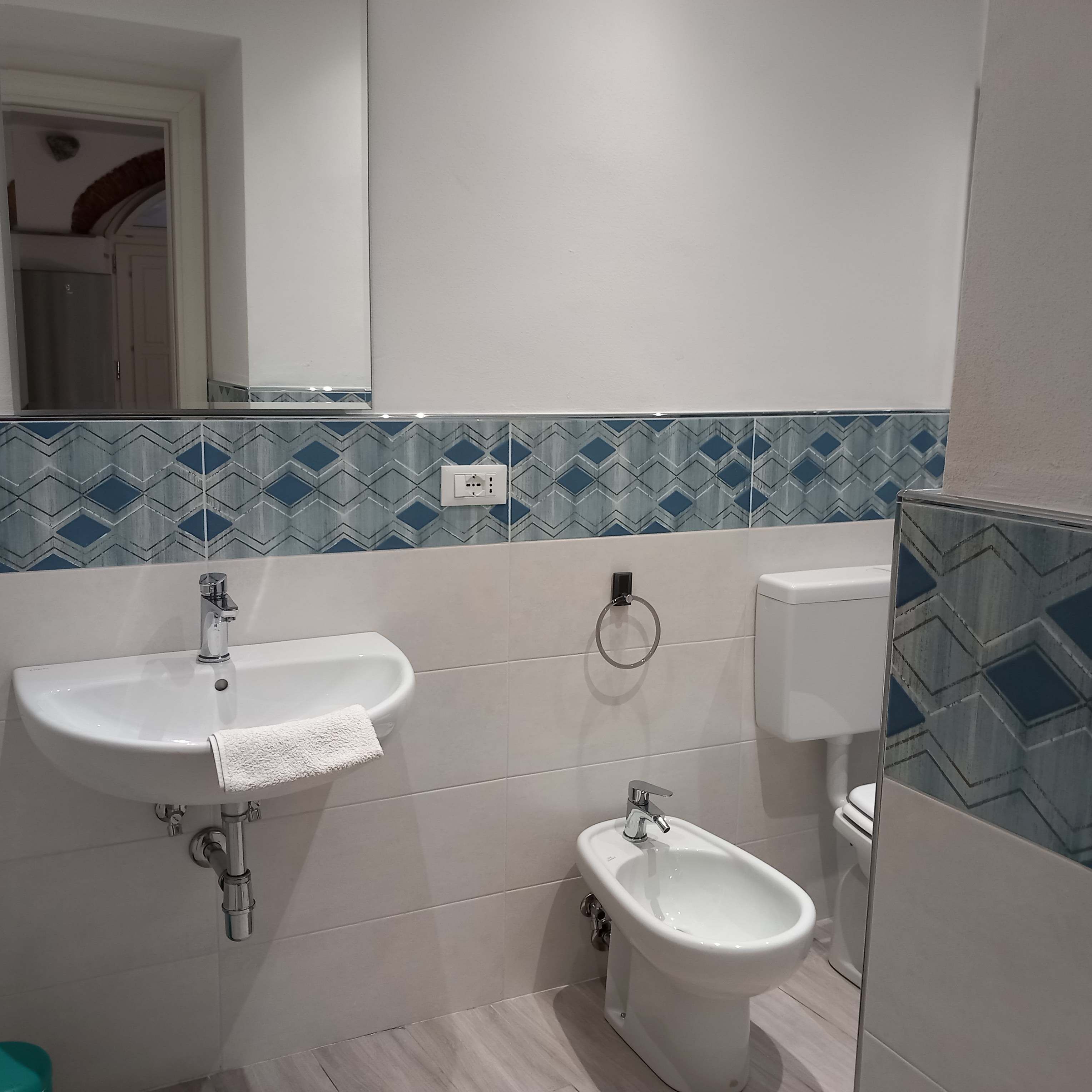 Een badkamer met een wit wastafel, een bidet en een toilet. Boven het wastafel hangt een spiegel en de spatwand is betegeld met een blauw patroon.