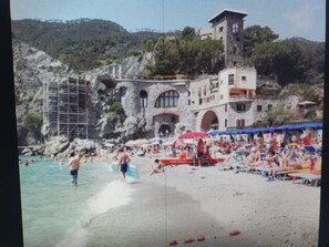 A beach with people, a building with arches, and a tower. - Casa Isabella Monterosso (Monterosso al Mare)