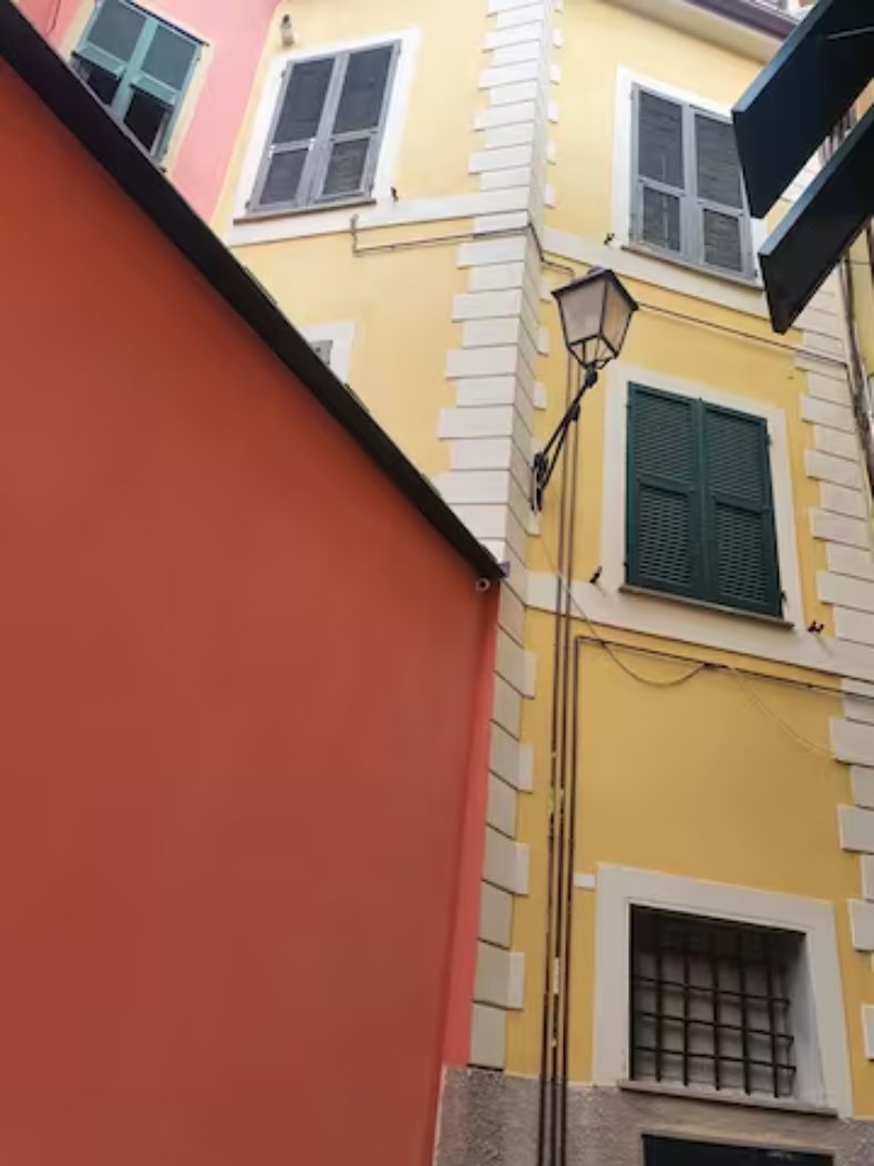 A narrow street with a yellow building featuring white window frames and shutters, a red wall, and a street lamp.