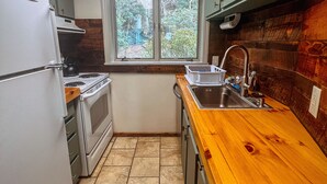 Een kleine keuken met een witte koelkast, fornuis en gootsteen. Boven de gootsteen bevindt zich een raam, en er zijn houten kastjes.