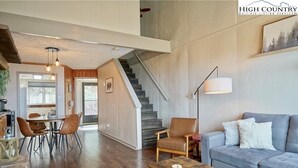 Een moderne woonkamer met een grijze bank, een houten salontafel en een eethoek met een houten tafel en stoelen. Een trap met een houten leuning leidt naar een hoger niveau.