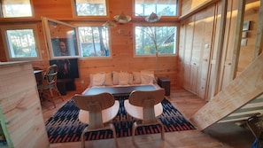 Quarto de cabana de madeira com cama, duas cadeiras, uma TV e janelas amplas.
